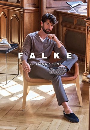 Gray v-neck sweater, tailored gray trousers, and blue slippers worn by a seated man in a wooden room with natural light. Accents include a black side table.
