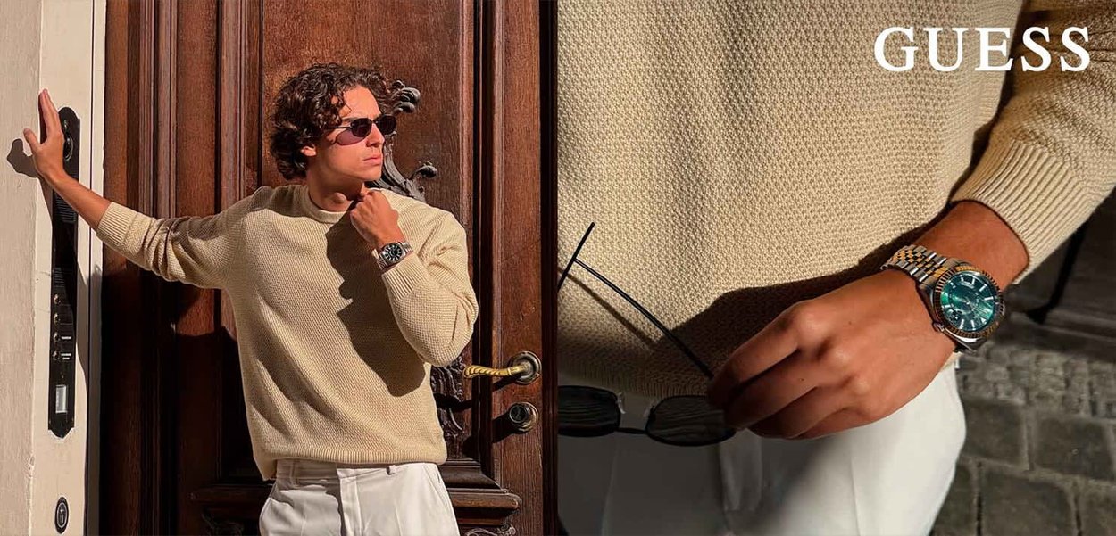 Beige gebreide trui met een textuurpatroon, gedragen met witte broeken. Een zilveren horloge met een groene wijzerplaat en een zonnebril in de hand.