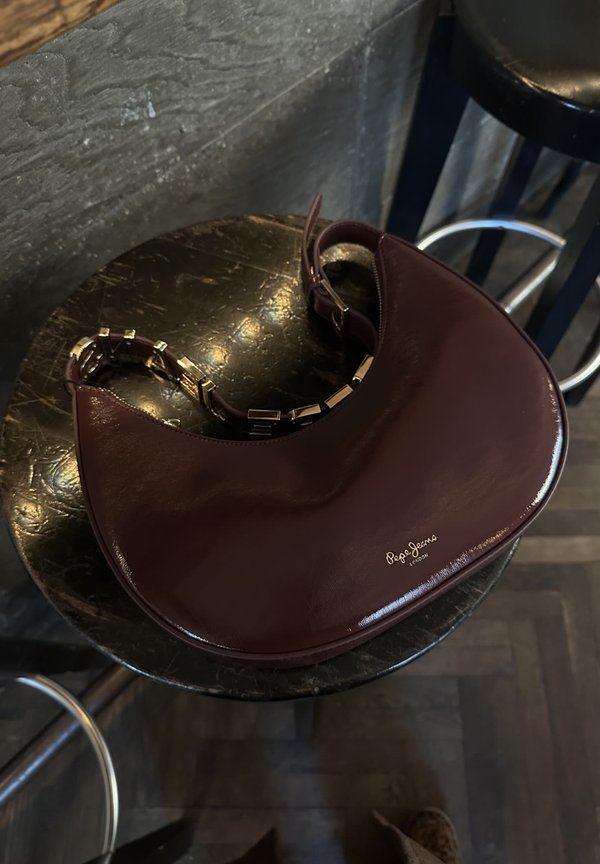 Bolso de mano de charol burdeos con forma curva, detalle de cadena dorada y logo grabado, descansando sobre una superficie de mesa redonda oscura.