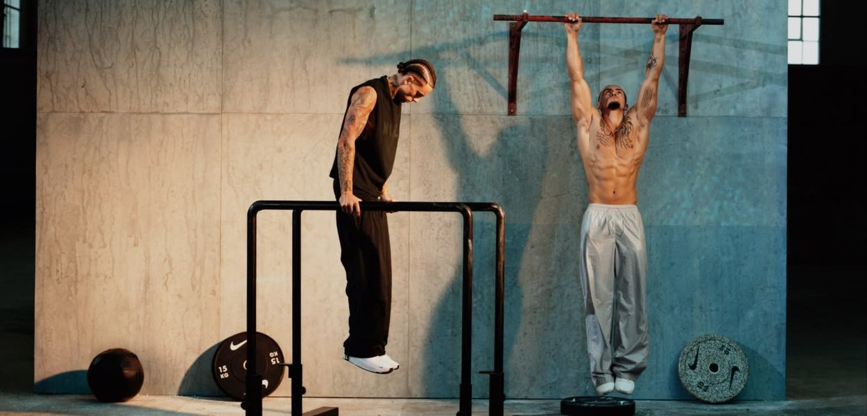 Dos hombres tatuados entrenando en interiores: uno haciendo fondos en barras paralelas, el otro realizando dominadas en una barra montada en la pared.