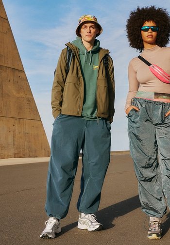 Un homme porte un sweat à capuche vert et une veste khaki, associés à un pantalon teal ample. Une femme en haut rose et pantalon gris brillant se tient à proximité, portant des lunettes de soleil.