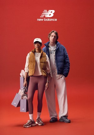 Due modelli posano davanti a uno sfondo rosso. Uno indossa un gilet imbottito color senape, leggings rosa e scarpe da ginnastica marroni, mentre l'altro è con una giacca a vento blu e pantaloni della tuta grigi. Entrambi tengono in mano delle buste della spesa.