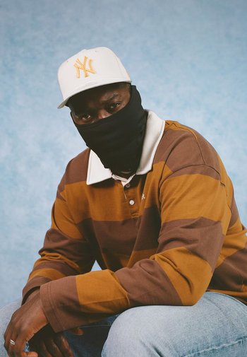 Bruin en geel gestreepte langemouwen overhemd, zwarte gezichtsmasker en witte baseballpet met geel logo. Lichtblauwe achtergrond.