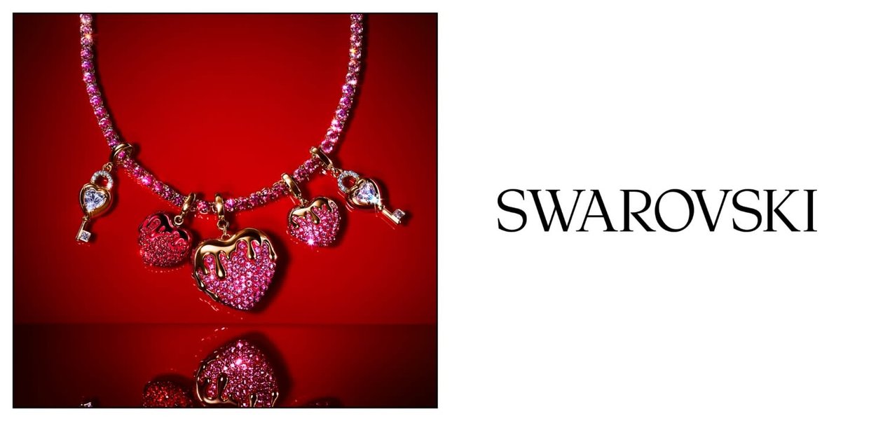 Różowy kryształowy naszyjnik z zawieszkami w kształcie serca i klucza na czerwonym tle, logo Swarovski na białym tle po prawej stronie.