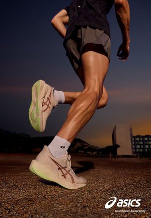 Läufer mit beigen Asics-Schuhen und weißen Socken, der beim Mittelschritt auf einer strukturierten Oberfläche in der Dämmerung mit Gebäuden im Hintergrund aufgenommen wurde.