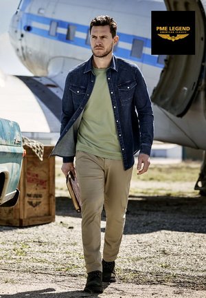 Denim shirt, green t-shirt underneath, beige pants; man walking with a book, vintage aircraft and blue truck in background.