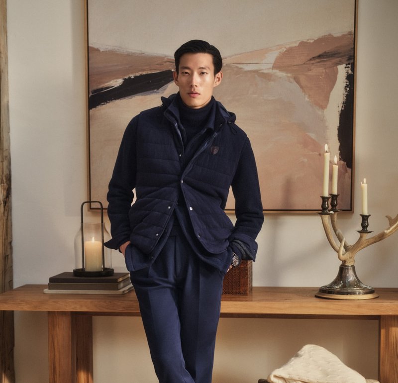 Mann in navy-blauem, geschichtetem Outfit lehnt an einem Holztisch mit brennenden Kerzen und einem abstrakten Gemälde in einem warmtonigen Raum.