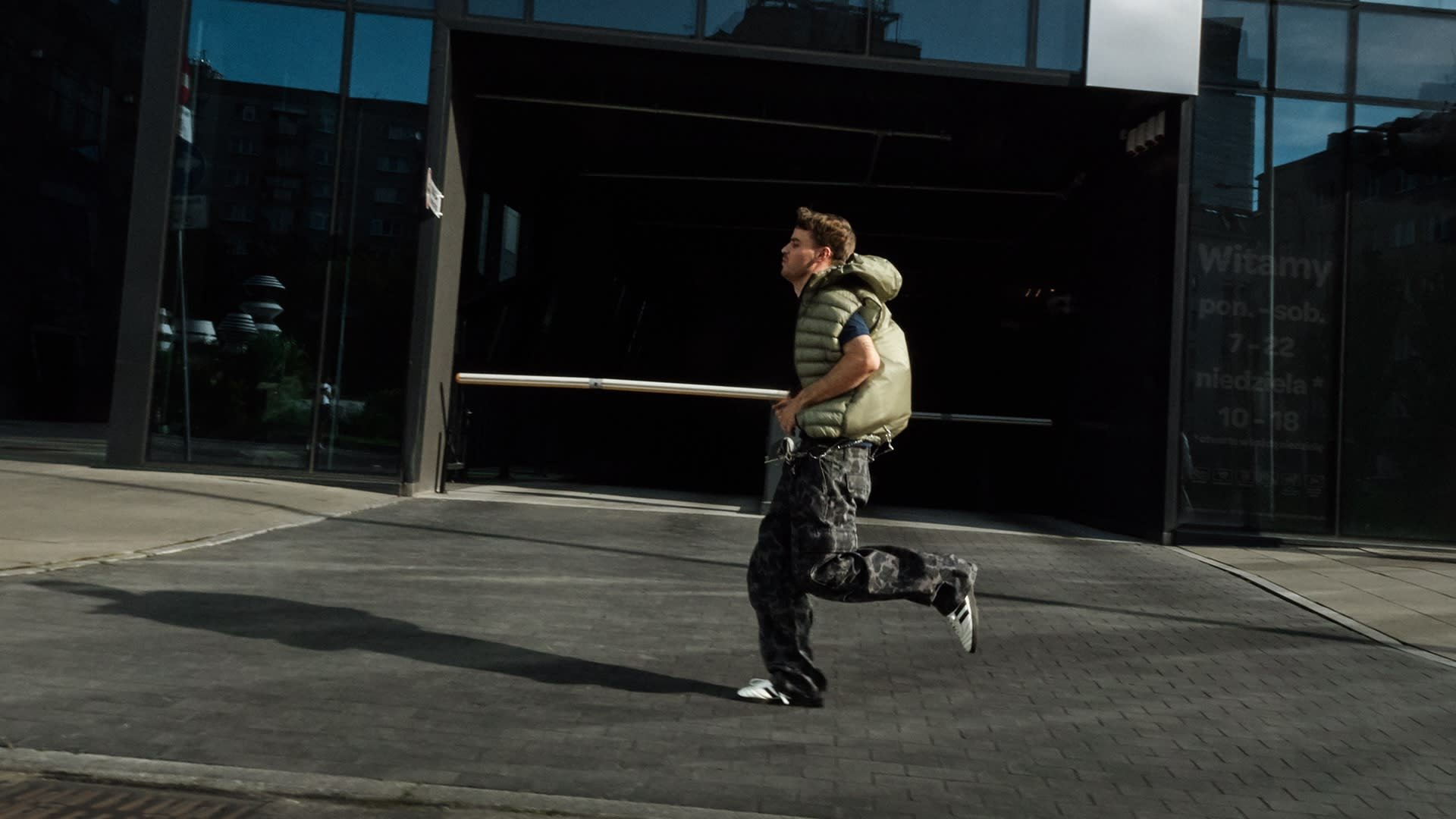 Gevoerde olijfgroene vest, camouflagebroek en zwarte schoenen; persoon joggend op een verharde wandelweg voor een glazen gebouw.