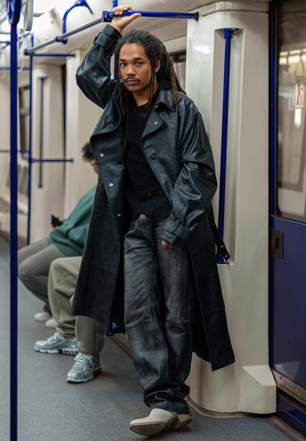 Cappotto lungo di pelle nera, maglione nero a coste, pantaloni di pelle grigi, scarpe crema e capelli testurizzati. Interno della metro con pali blu.