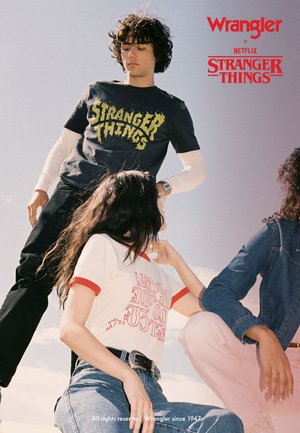 T-shirt en coton noir avec l'inscription "STRANGER THINGS" en vert, rouge et blanc dans un style graphique vintage, bordure rouge, et veste bleue. Cadre décontracté.