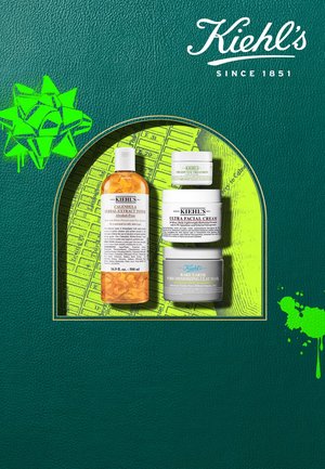 Kiehl's Hautpflegeprodukte auf einem grünen Lederhintergrund: Bernstein-Tonerflasche, weiße Gesichtscreme und graue Tonmaske mit klaren Etiketten.