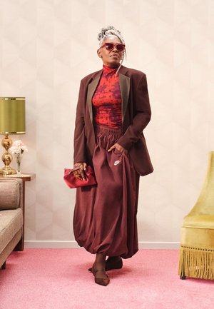 Gonna oversize color granata abbinata a una maglietta rossa con motivo e blazer marrone. Accessori includono una pochette rossa e occhiali da sole tondi. Sfondo testurizzato.