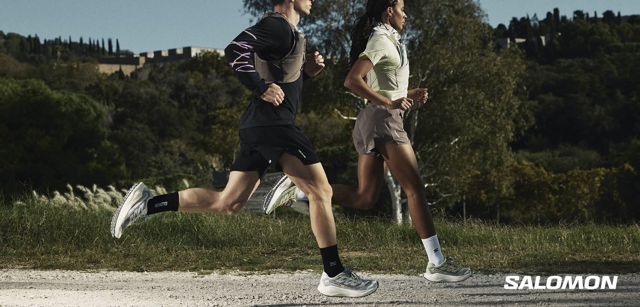 Deux coureurs, un homme et une femme, joggent côte à côte sur un sentier de terre entouré d'herbe et d'arbres en plein jour.