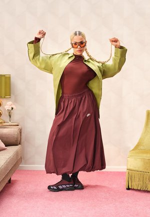 Camicia oversize in raso verde, top aderente color prugna, gonna color prugna con logo Nike, sneakers nere, occhiali da sole, capelli intrecciati, sfondo con moquette rosa.