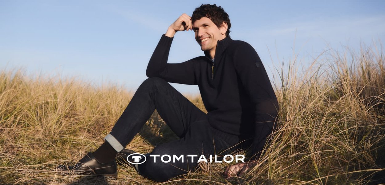Mężczyzna w czarnym swetrze i ciemnych dżinsach siedzi i uśmiecha się na suchej trawie pod bezchmurnym niebem, wyświetlone logo Tom Tailor.