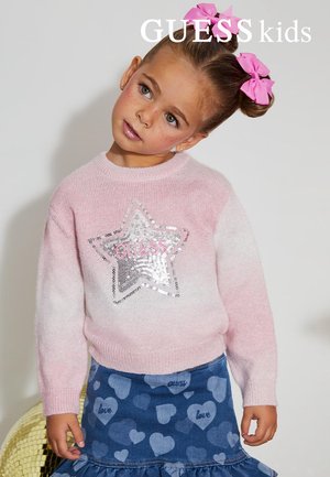 Jeune fille avec des nœuds roses, portant un pull rose avec une étoile argentée et une jupe bleue à motifs de cœurs, inclinant la tête.