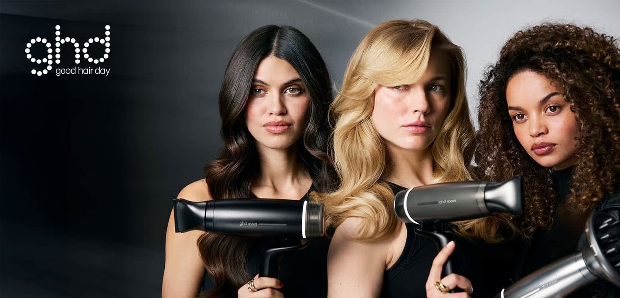 Tre kvinder med stylet hår holder sorte ghd-hårtørrere og står mod en neutral baggrund med ghd-logoet i øverste venstre hjørne.