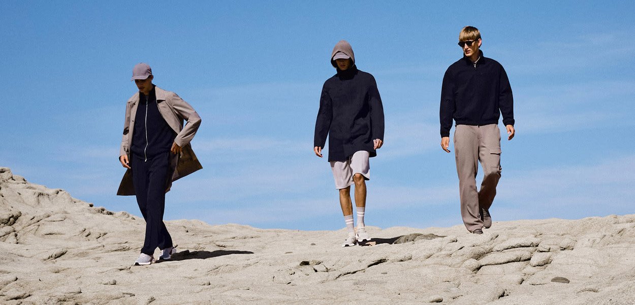 Tre uomini camminano su un terreno roccioso sotto un cielo azzurro limpido, indossando abiti casual a strati con cappelli e occhiali da sole.