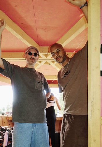 Zwei Männer stehen in einer Holzstruktur mit einer pinkfarbenen Decke. Der eine trägt eine Sonnenbrille und ein graues Shirt; der andere hat ein braunes Shirt mit dem Aufdruck "FORMER".