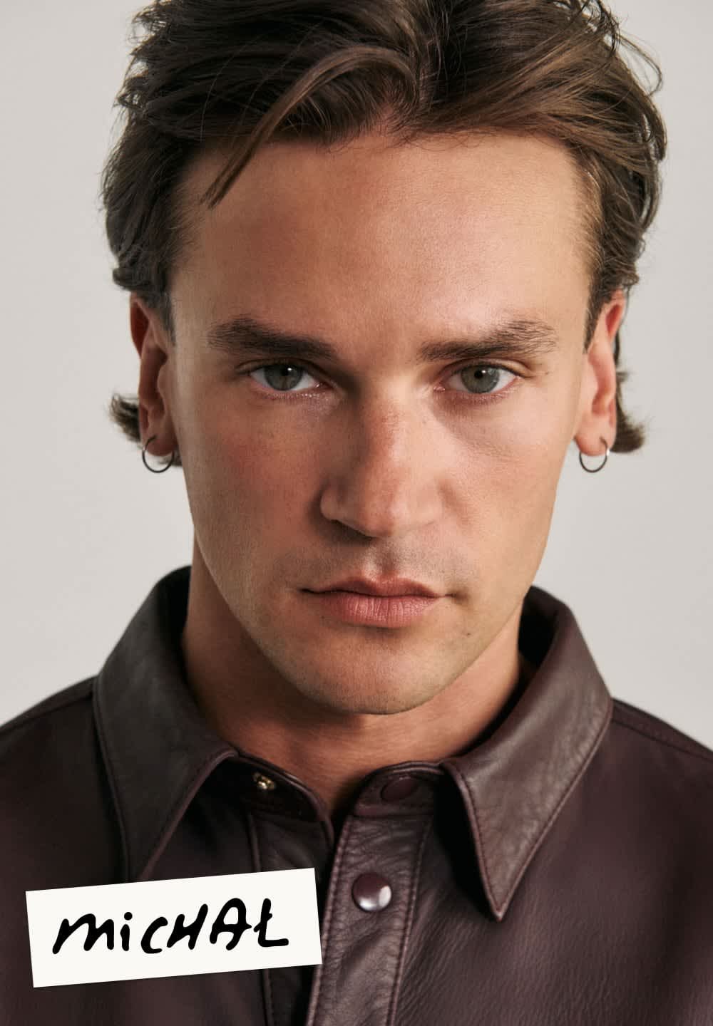 Brązowa skórzana kurtka z kołnierzem, zapinana na guziki, noszona przez osobę z krótkimi, lekko falującymi włosami i kolczykiem w kształcie kółka.