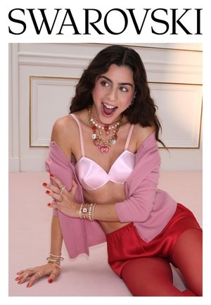 Vrouw met roze beha, vest, rode korte broek en gelaagde kettingen en armbanden, lachend met open mond tegen een witte paneelmuur.