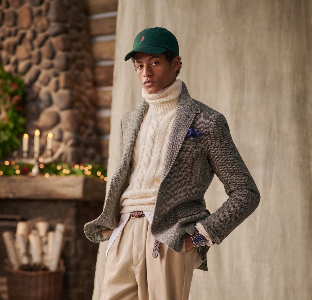 Blazer grigio a spina di pesce sopra un dolcevita bianco a trecce, pantaloni beige, cappello verde, fazzoletto da taschino blu e orologio, su uno sfondo rustico.