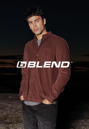 Braunes Cordhemd mit Knöpfen und lockerem Sitz, mit Brusttasche und sichtbaren Nähten, getragen über einem grauen T-Shirt und schwarzen Jeans.