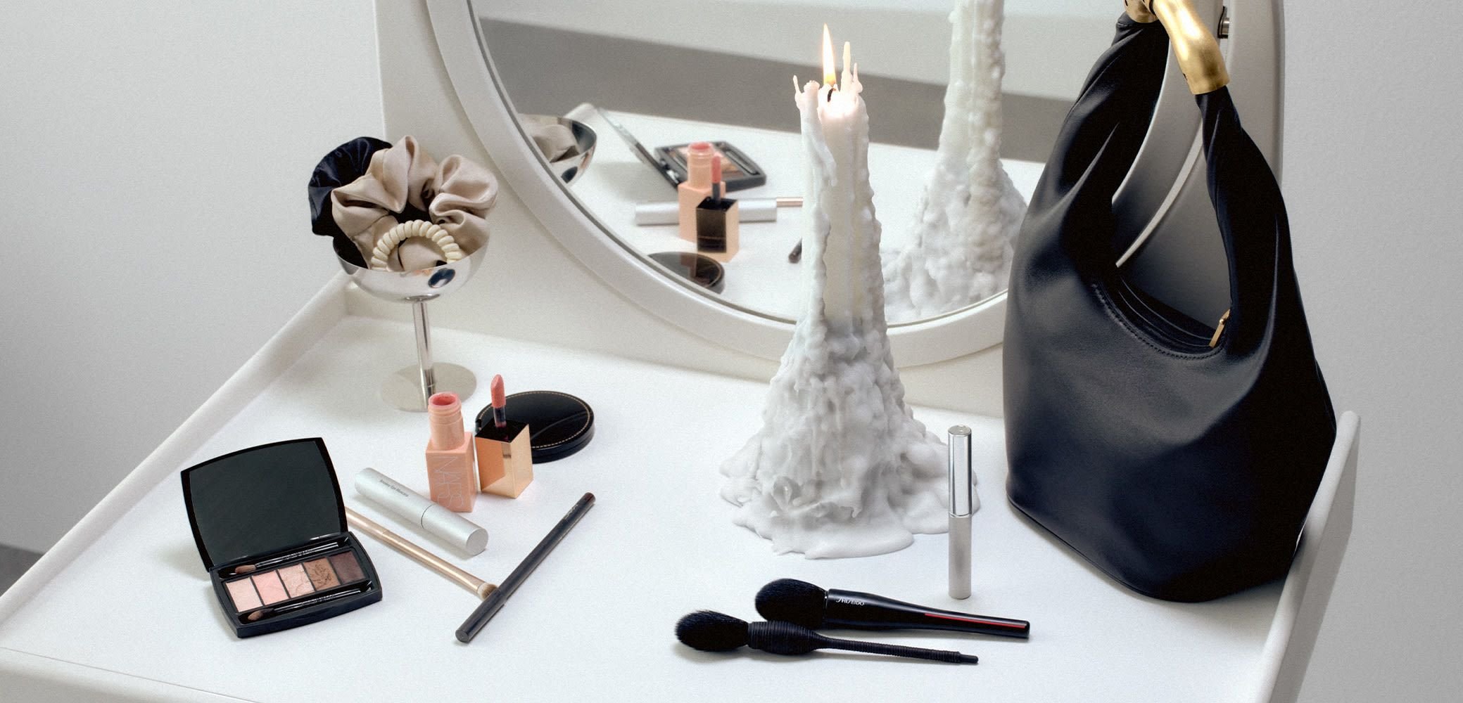 Zwarte handtas met gouden hardware, make-upproducten, borstels, een kaars en accessoires op een witte tafel naast een ronde spiegel.