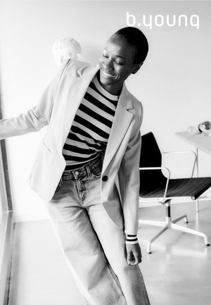 Blazer sartoriale grigio chiaro su una camicia a righe bianca e nera, abbinato a jeans di denim blu a vita alta. Tessuto liscio, vestibilità moderna.