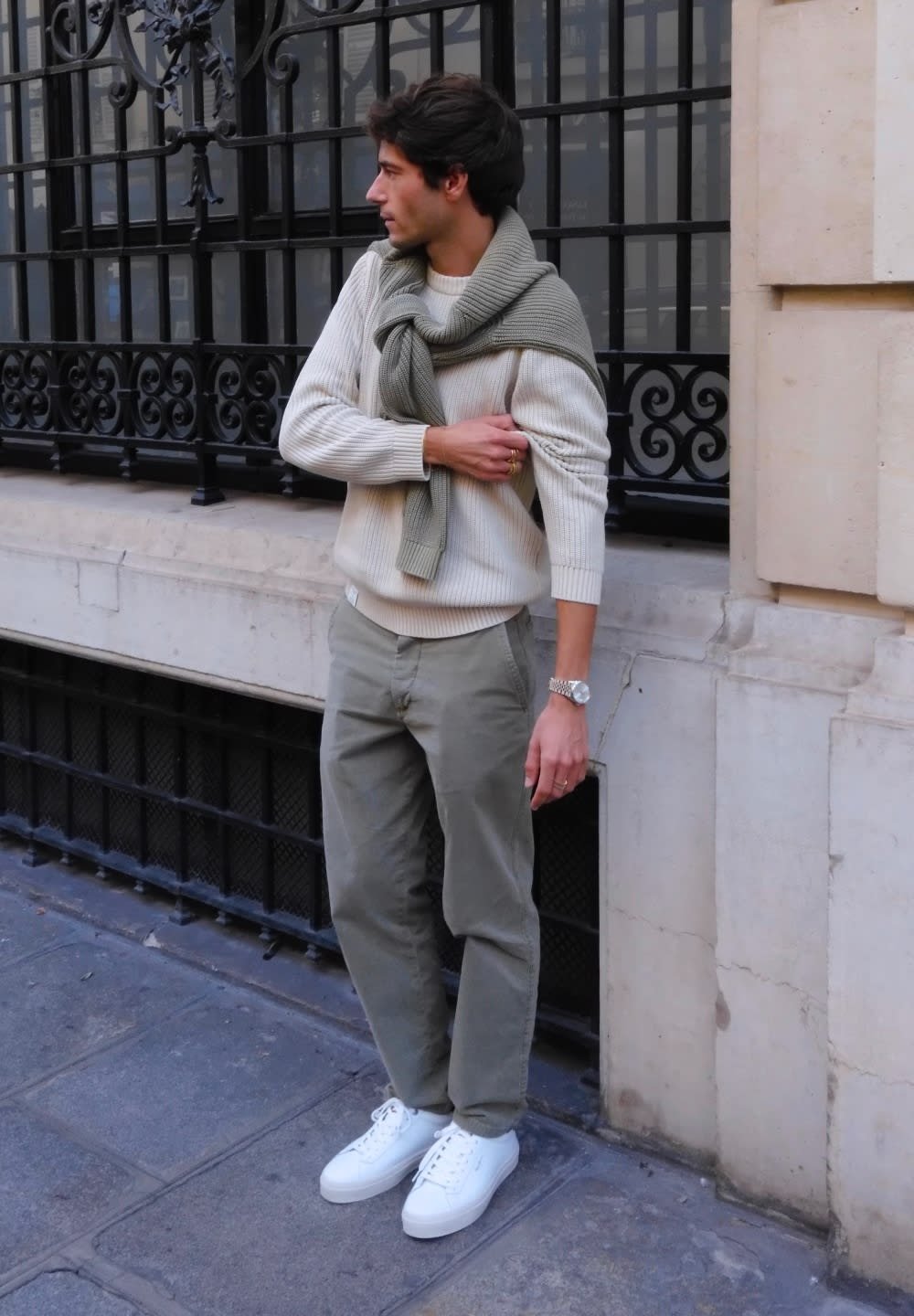 Suéter blanco de canalé con una bufanda de punto verde claro, pantalones verdes oliva y zapatillas blancas. Modelo de pie contra una fachada de edificio.