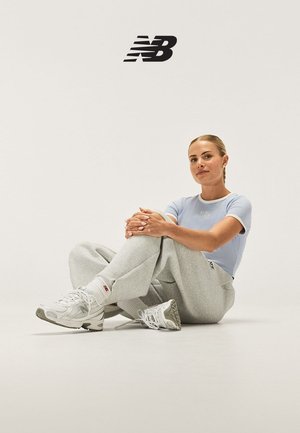 Femme assise par terre portant un t-shirt New Balance bleu clair, un pantalon de survêtement gris, des baskets blanches New Balance, sous un logo NB noir sur fond uni.