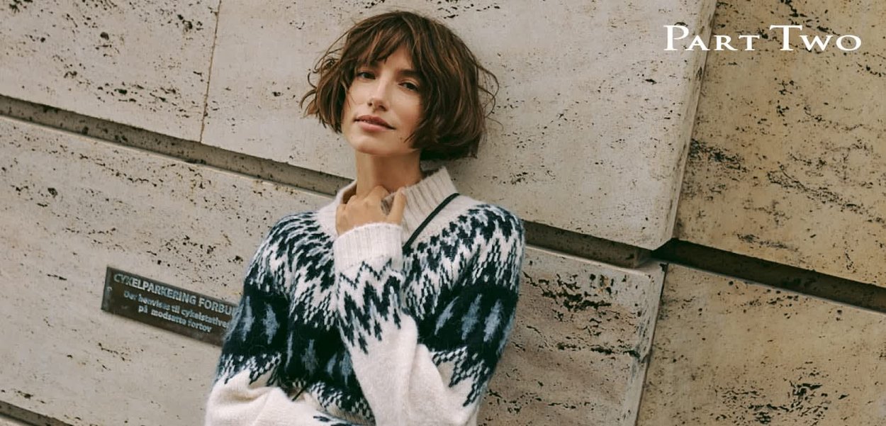 Strickpullover in Creme mit dunkelblauen, geometrischen Mustern, ausgestattet mit V-Ausschnitt und gerippten Bündchen. Vor einer strukturierten Steinmauer inszeniert.