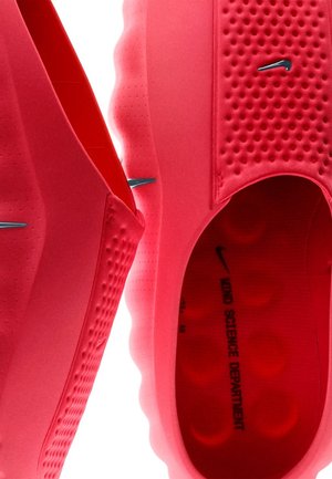 Zapatos rosas brillantes sin cordones con material liso y flexible, lados texturizados y patrones circulares en la suela. Marca Nike en azul.