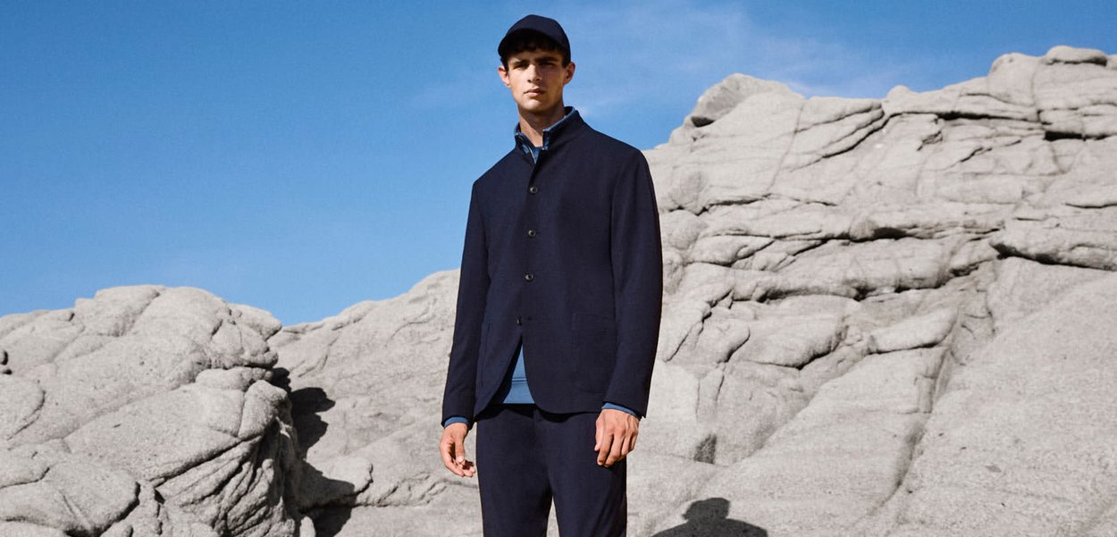 Jonge man in donkerblauw pak en pet staat op rotsachtig terrein onder een heldere blauwe lucht.