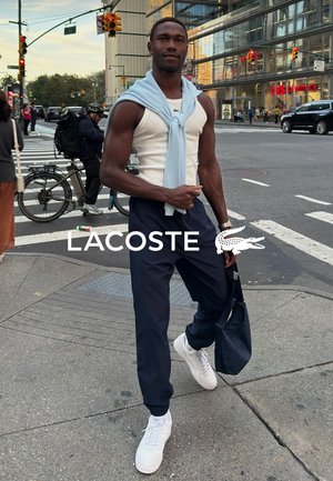 Top blanco de tirantes combinado con un suéter liviano azul claro, pantalones azul marino y zapatillas blancas. La persona sostiene un bolso azul marino y está de pie en la calle.