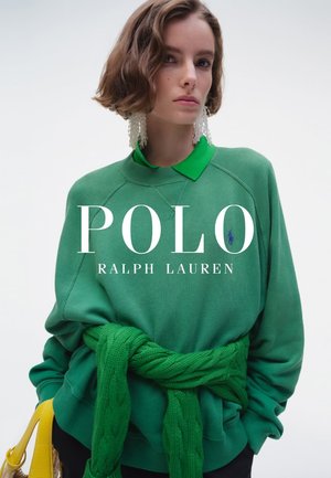 Joven mujer con sudadera verde Polo Ralph Lauren con jersey a juego atado en la cintura y grandes pendientes blancos, sosteniendo una bolsa amarilla.