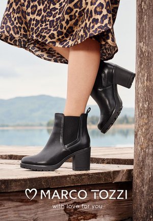 Black ankle boots with a chunky heel, elastic side panels, smooth leather texture, and a low-profile sole, set against a wooden background.