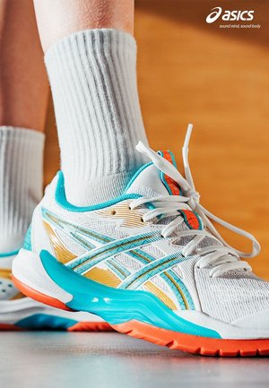 White and turquoise ASICS running shoes with orange accents worn with white socks on a wooden floor, ASICS logo in top right corner.