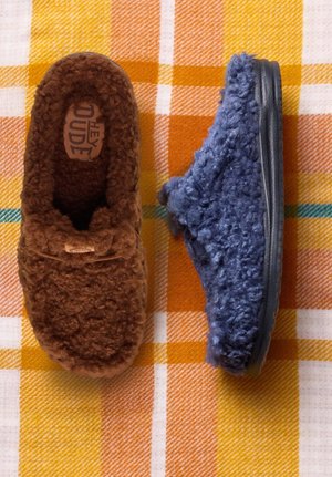 Zwei Paar flauschige Hausschuhe: eines braun mit weichem, strukturiertem Obermaterial, das andere blau mit ähnlicher Struktur und dunkler Sohle, auf einem karierten Hintergrund platziert.