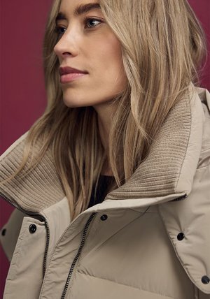 Beige dynejakke med høj krave og ribbet detaljer, med lynlås foran og trykknapper, båret mod en mørkerød baggrund.