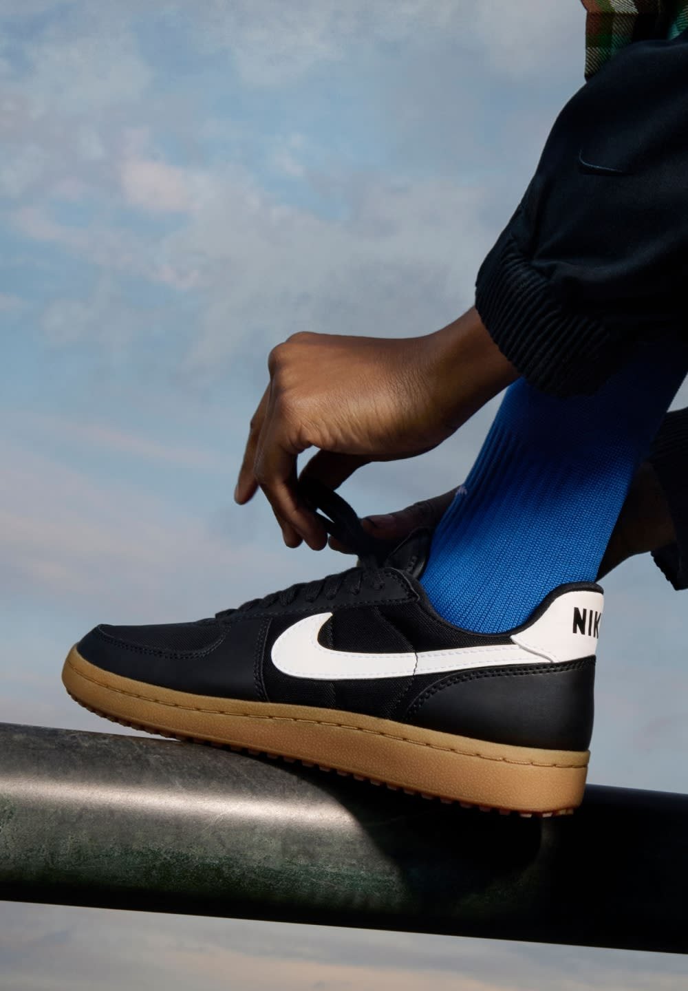 Nike neri con swoosh bianco e suola in gomma, indossati con una calza blu. Primo piano di una mano che allaccia la scarpa su un corrimano di metallo.