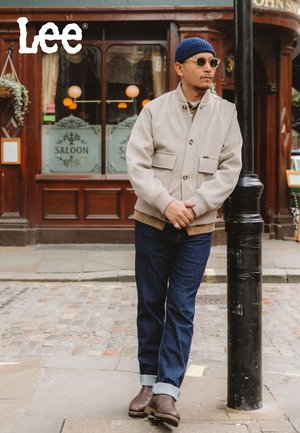 Beige Jacke mit Knöpfen und Fronttaschen, dunkelblaue Jeans, braune Stiefeletten und eine blaue Strickmütze, stehend bei einer Straßenlaterne.