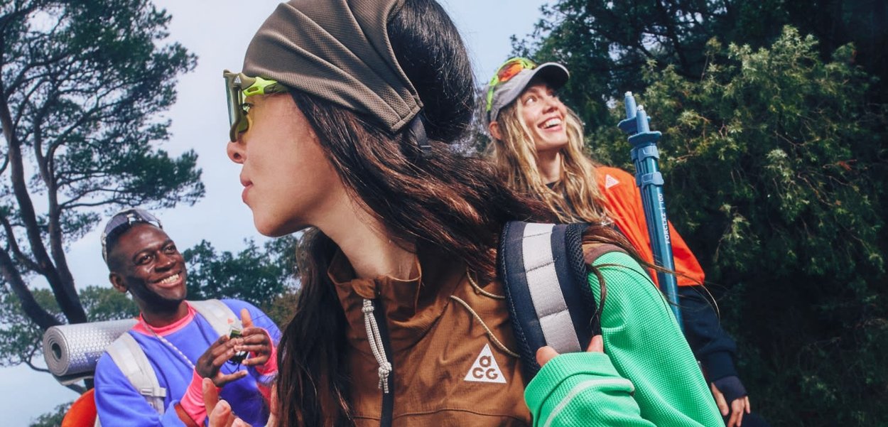 Tres excursionistas con ropa y mochilas de senderismo sonríen y conversan en un área boscosa con pinos.