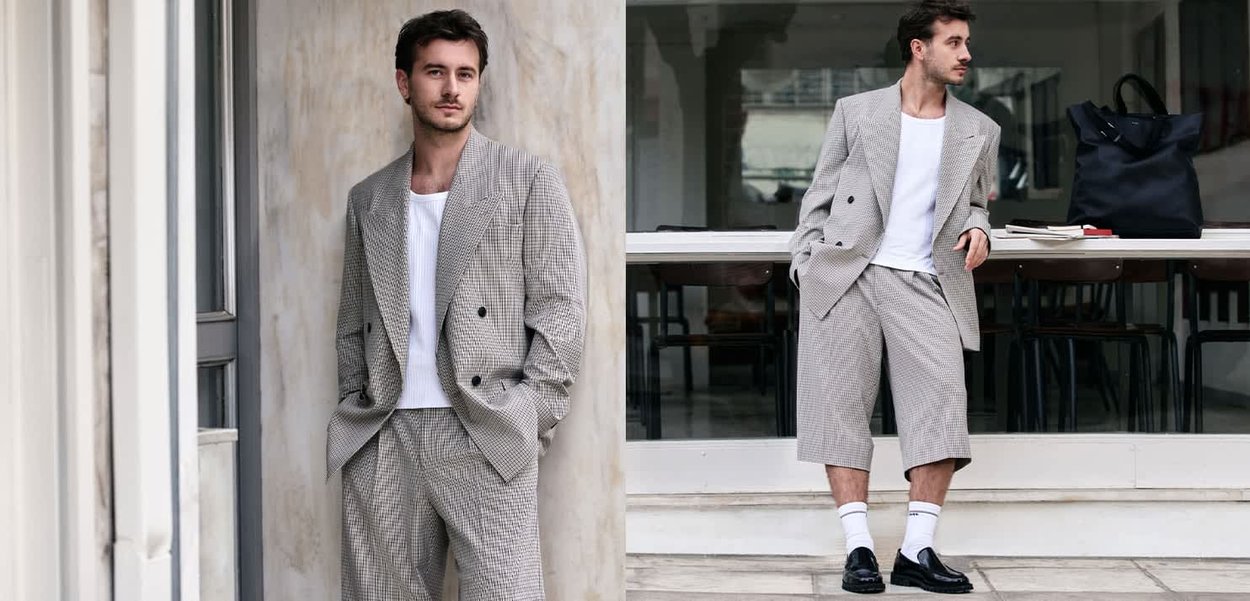 Hombre vestido con blazer a cuadros gris y pantalones cortos con camiseta blanca y mocasines negros, de pie y apoyado contra una pared y escalones fuera de un edificio.
