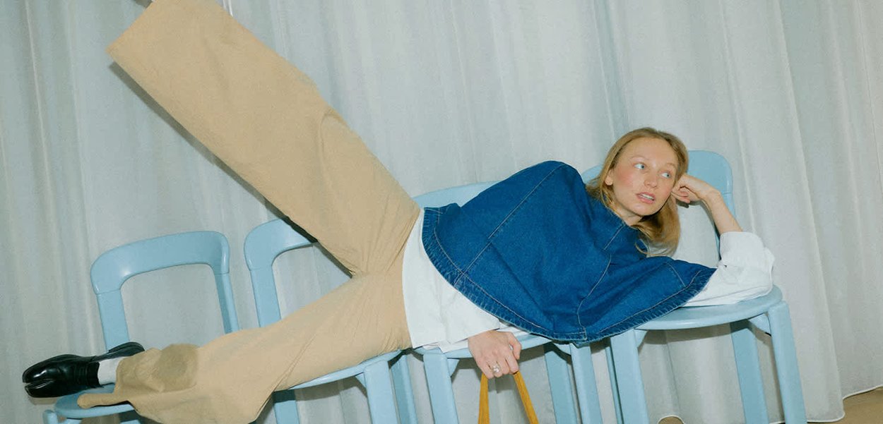 Frau in beigen Hosen und blauem Denim-Oberteil liegt seitlich über drei blaue Stühle vor weißem Vorhang.