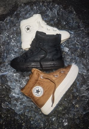 Drei hohe Converse All Star Sneakers in Weiß, Schwarz und Braun sind auf verstreuten Eisklumpen angeordnet.
