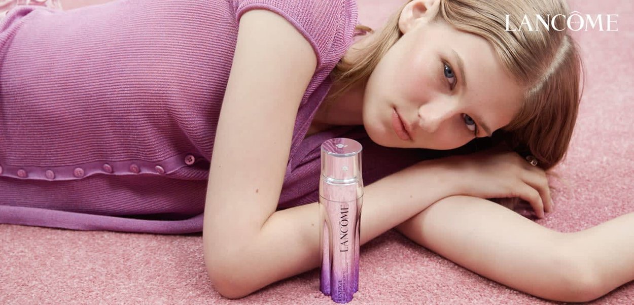 Lancôme-serum i en klar flaska med en lila gradientdesign, placerad på en mjuk rosa yta, bredvid en modell i en texturerad lila topp.