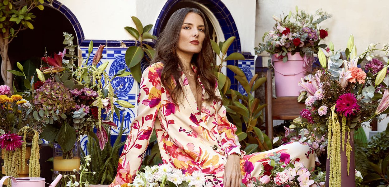 Mujer con traje floral sentada entre arreglos florales coloridos y plantas verdes contra un fondo de pared azul y blanca con azulejos.