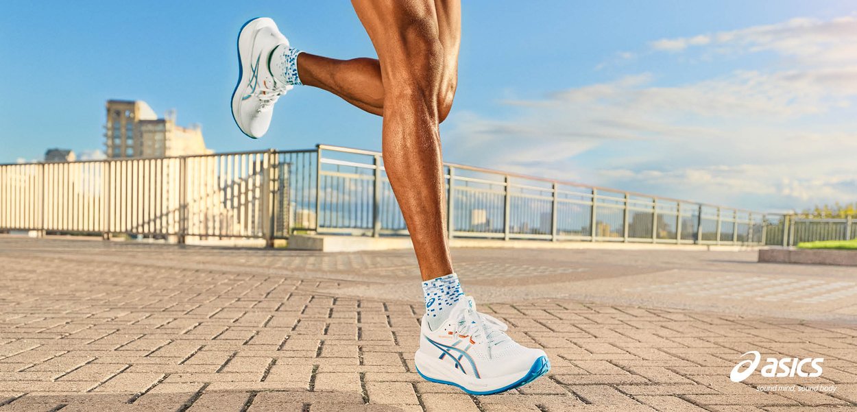 Getrainde benen van een hardloper in witte en blauwe Asics-schoenen op een geplaveid buitenspoor met leuningen en een heldere lucht op de achtergrond.