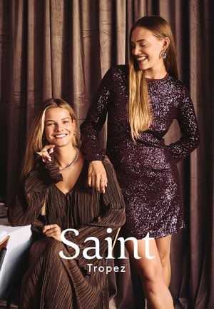 Two women pose together; one wears a form-fitting, purple sequined dress, the other a brown pleated blouse with ruffle sleeves.